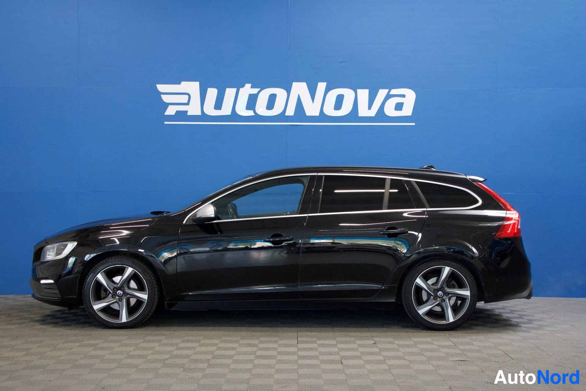 Sivulta: Musta Volvo V60 sivusta vasemmalla, näkyvissä koko vasen sivu ja edestä takaa, valkoiset vanteet, sininen seinä taustalla