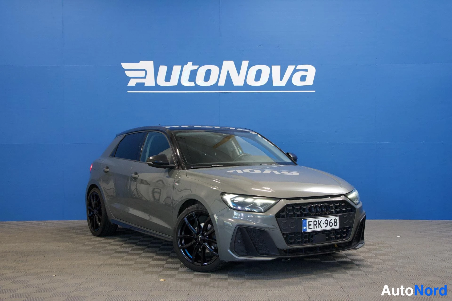 Etuviisto, nokka oikealle: Harmaa Audi A1 Sportback kuvattuna kolmen neljänneksen kulmasta vasemmalta, sinisen seinän edessä. Rekisterikilvessä