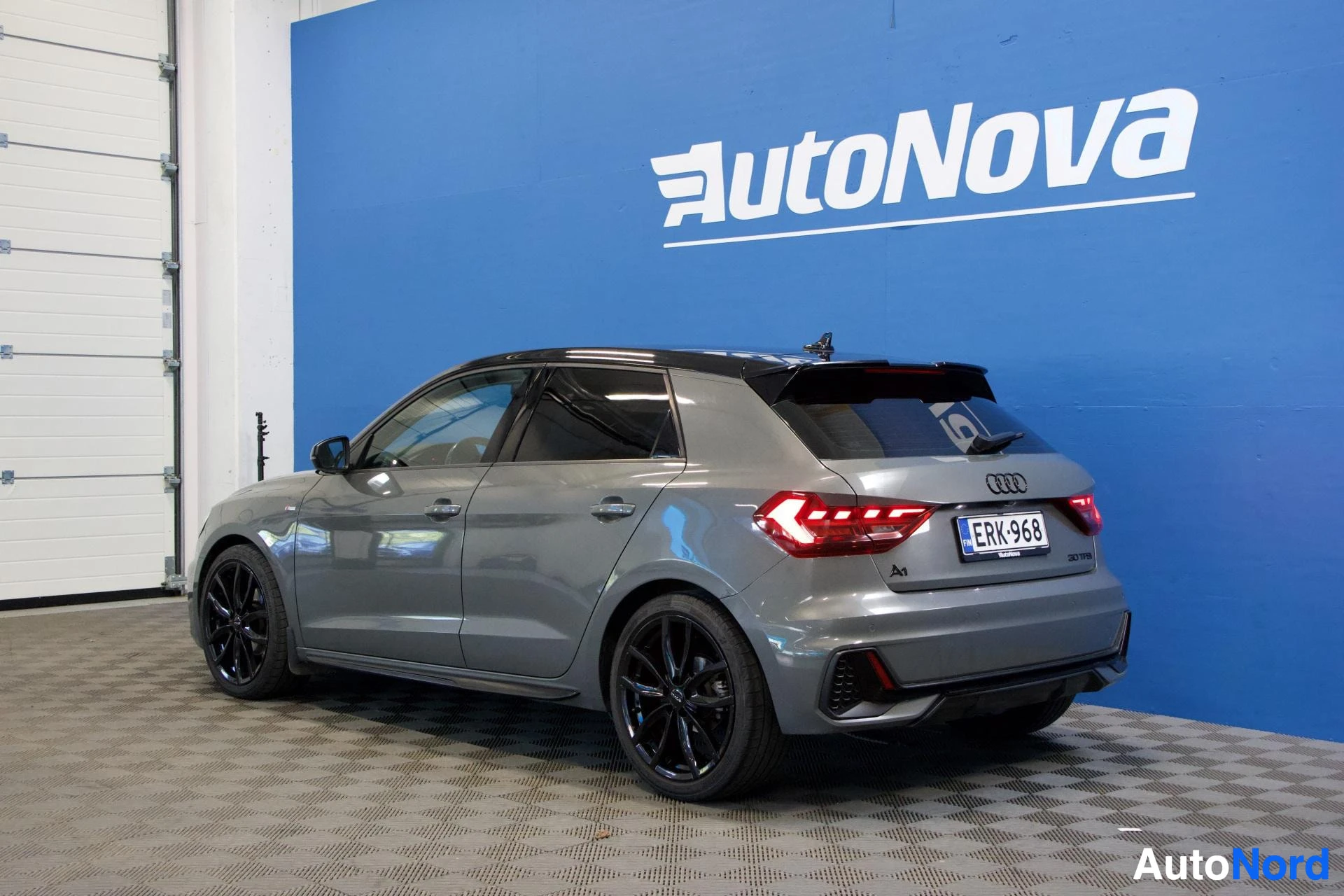 Takaviisto: Audi A1 Sportback takaa kolmelta kulmalta, ovi auki, rekisterinumero, harmaa väri, tummat vanteet, valkoinen seinä ja sininen tausta