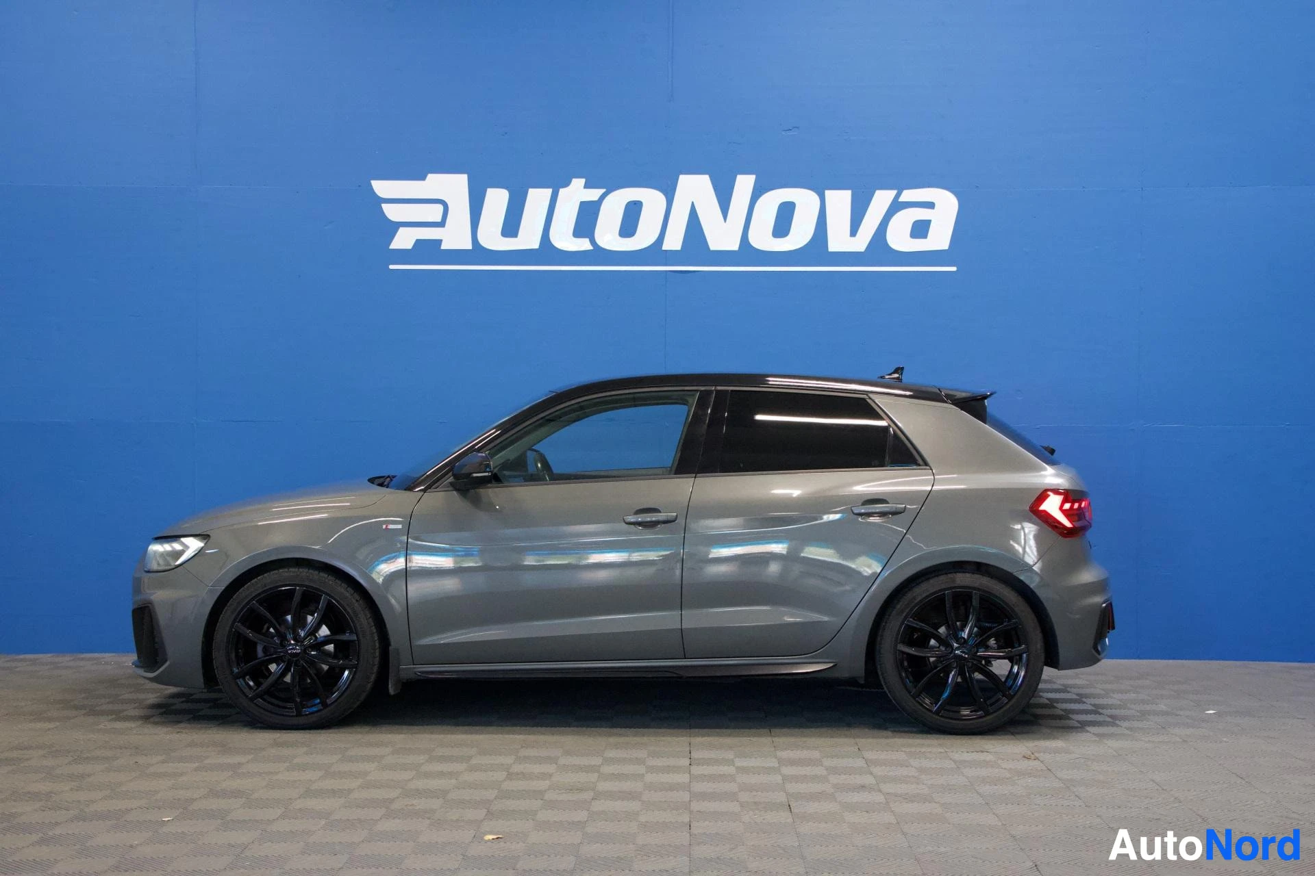Sivulta: Audi A1 Sportback sivusta vasemmalta, koko auto näkyy, tummat vanteet, harmaa väri, molemmat ovet auki, sininen tausta
