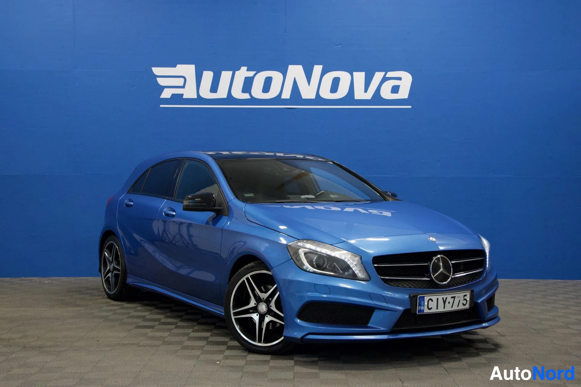 Etuviisto, nokka oikealle: Sininen Mercedes-Benz A -henkilöauto etuviistosta valokuvattuna sinisellä AutoNova-taustalla, rekisteritunnus.