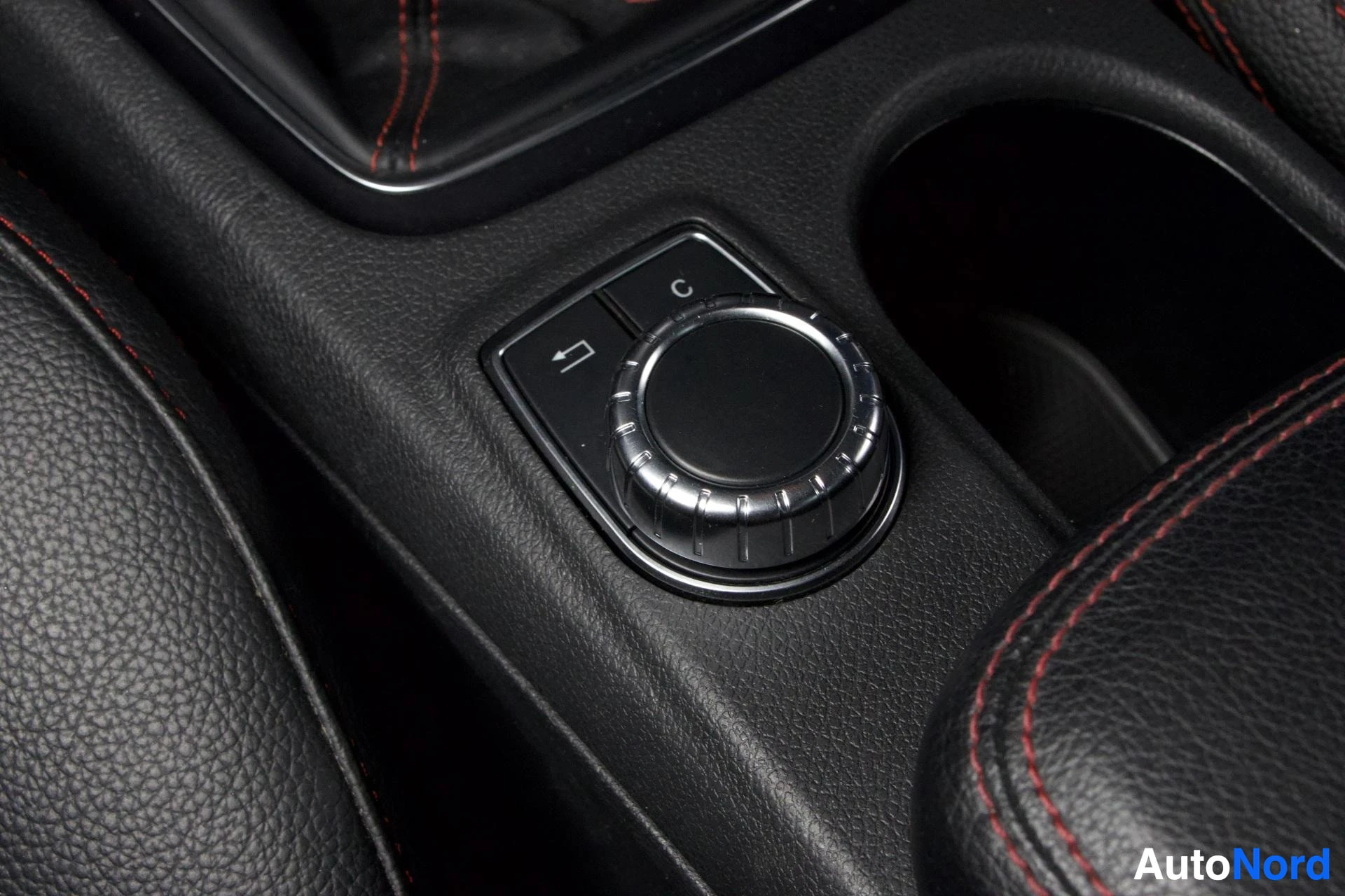Muu: Sisätilakuvan keskikonsolin Mercedes-Benz COMAND-ohjausnuppi merkki-merkinnöin C ja takaisinnuoli, punaisilla ompeluksilla.