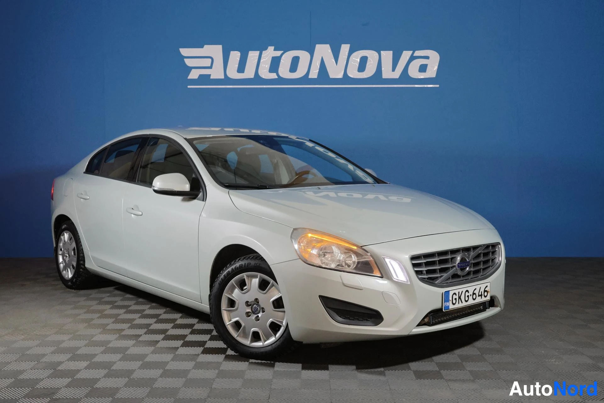 Etuviisto, nokka vasemmalle: Valkoinen Volvo S60 -sedan kuvattuna kolmen neljänneksen kulmasta vasemmalta, näkyy etuosa ja vasen sivu. Ajoneuvolla on valkoiset vanteet ja sininen t