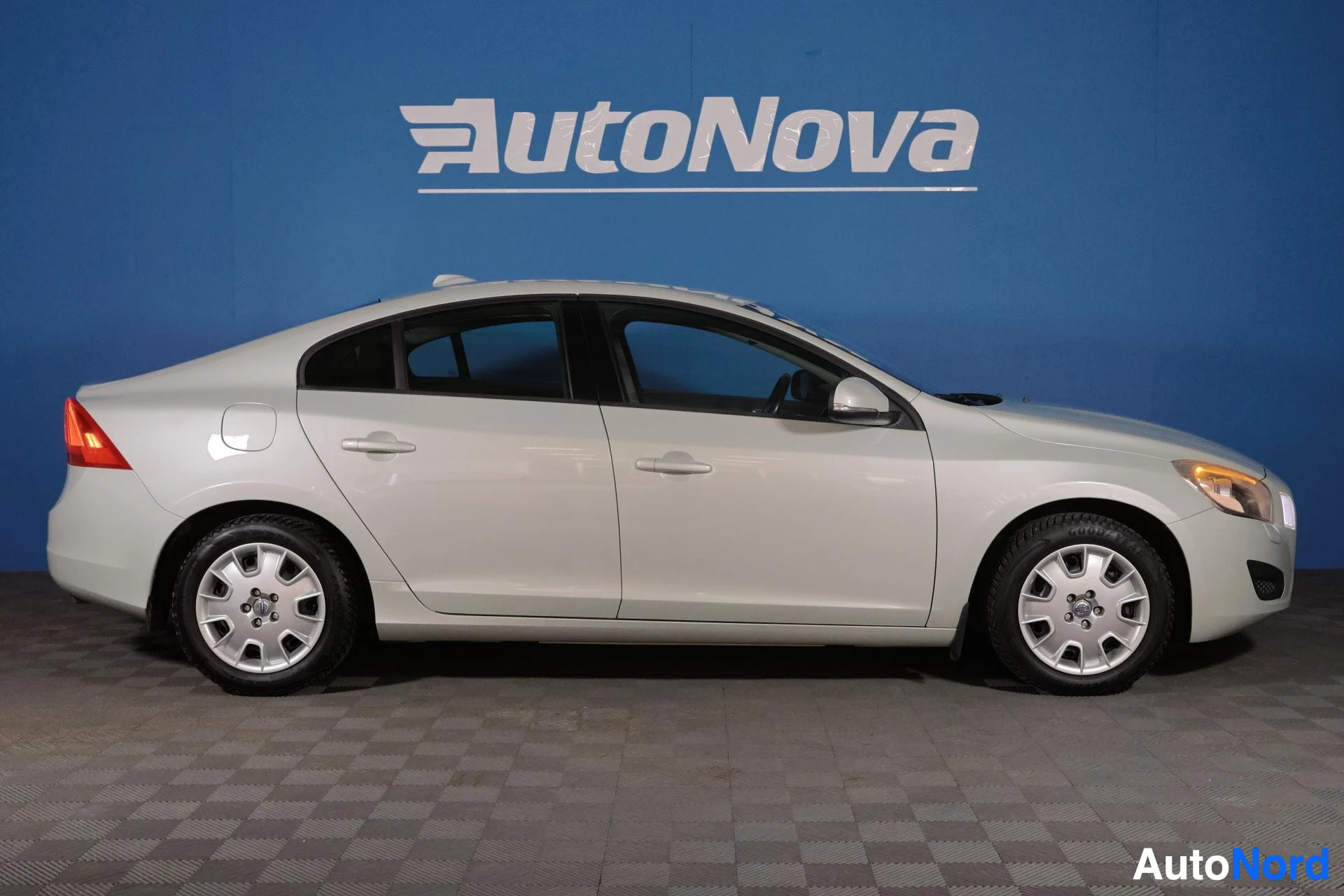 Sivulta: Valkoinen Volvo S60 -sedan sivusta, rekisteritunnusta ei näkyvissä, alumiiniset navakuvut, paikoittuneena sinisellä seinustalla.