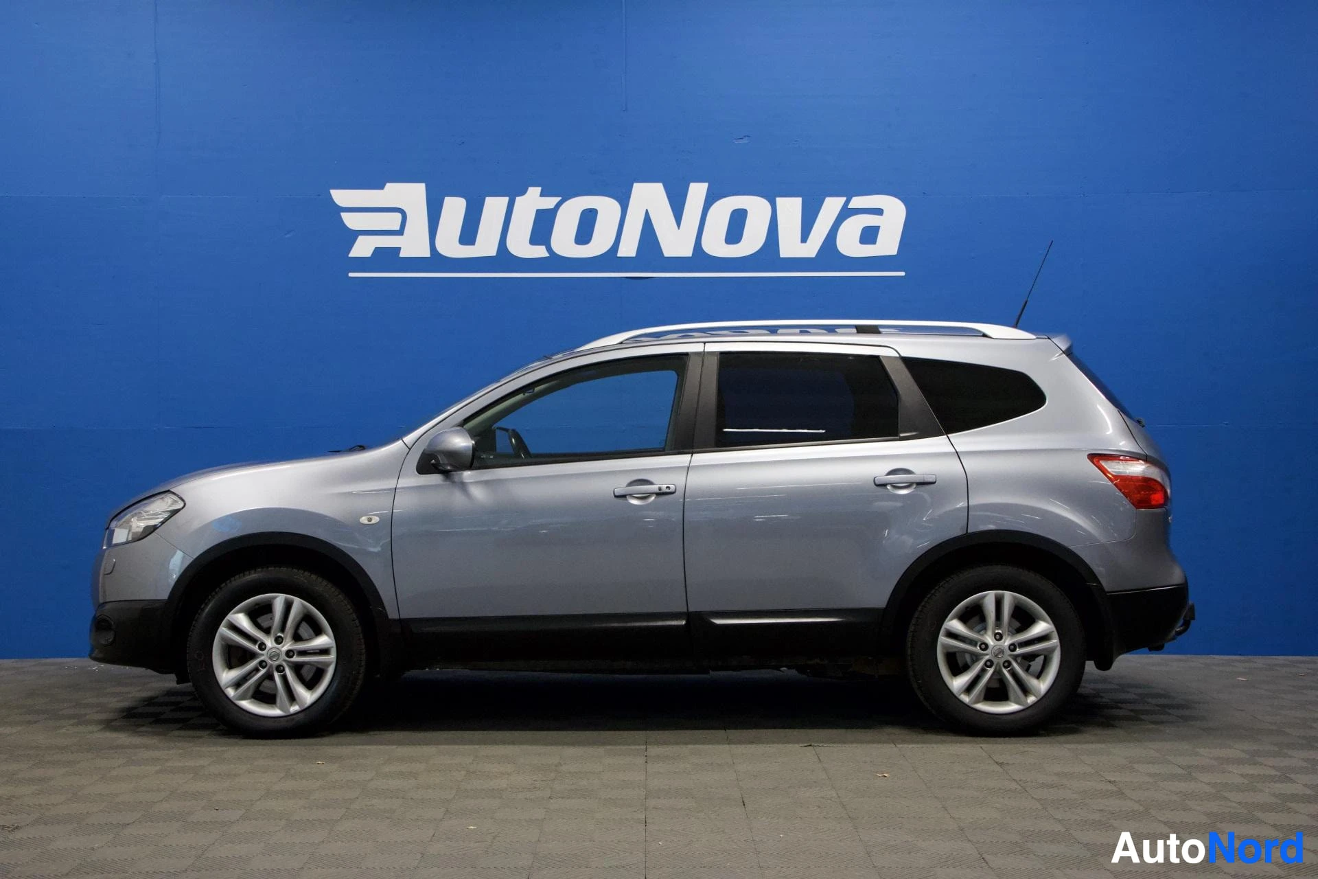 Sivulta: Hopeanvärinen Nissan Qashqai+2 SUV, kuvattu sivusta oikealle päin, sinisellä AutoNova-taustalla.