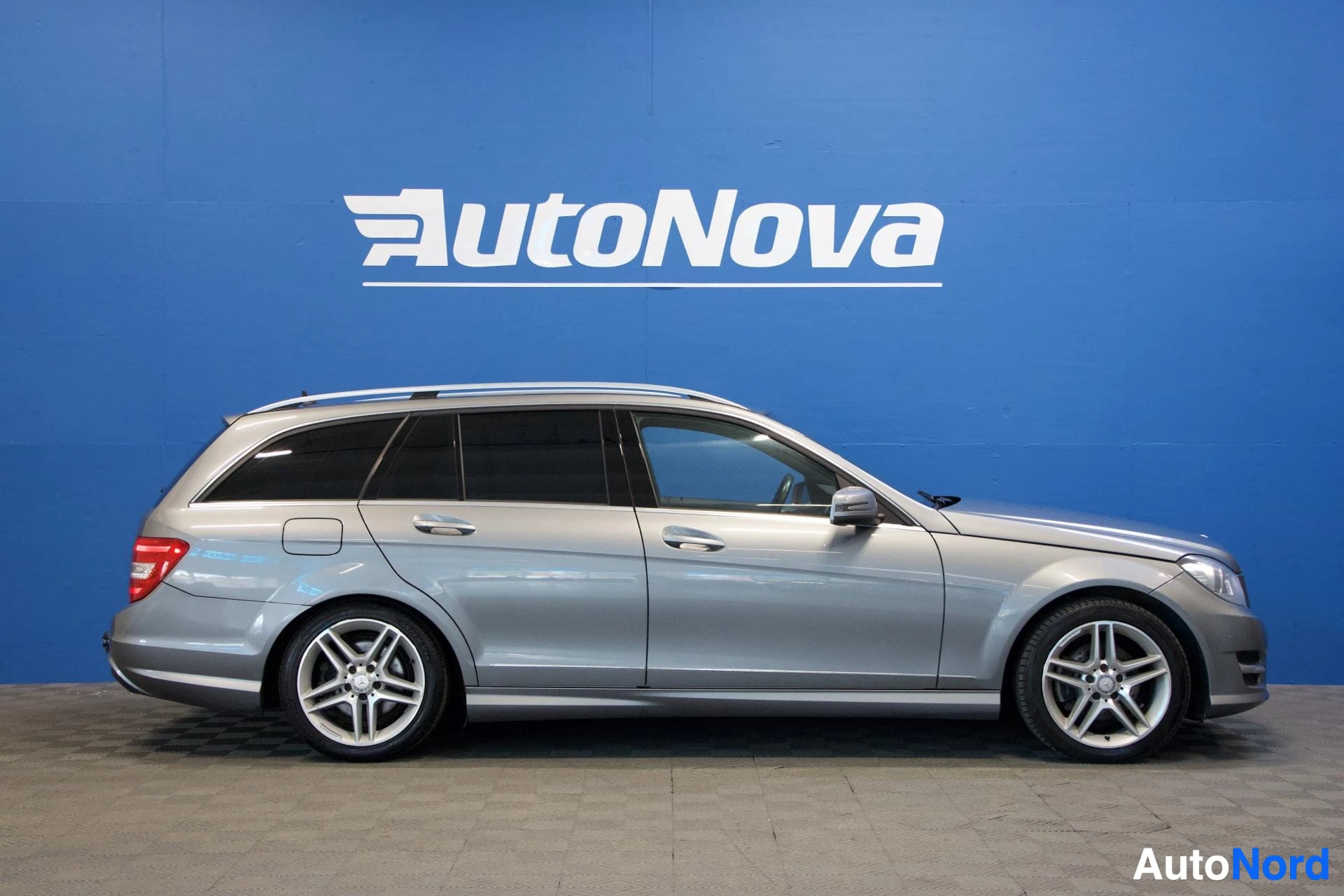 Sivulta: Hopea Mercedes-Benz farmari seisoo profiilinäkymässä sinisen autoNova-seinän edessä.