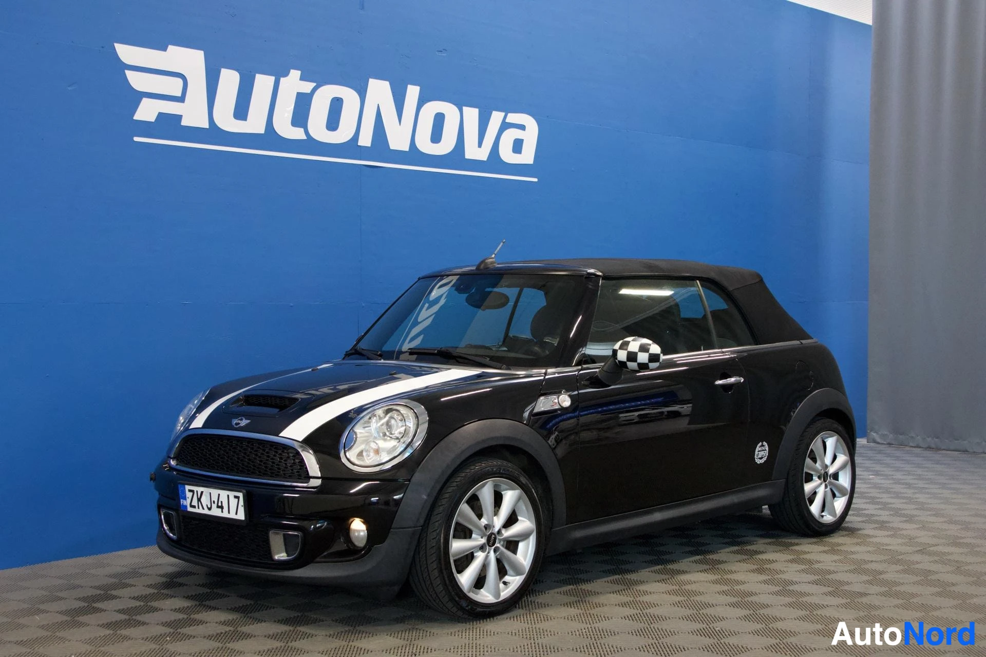 Etuviisto, nokka vasemmalle: Musta Mini Cooper avoauto edestä kolmen neljänneksen kulmasta katetussa tilassa, valkoinen keskijuova, rautatieaseman logo peilissä, valkoisen ja musta