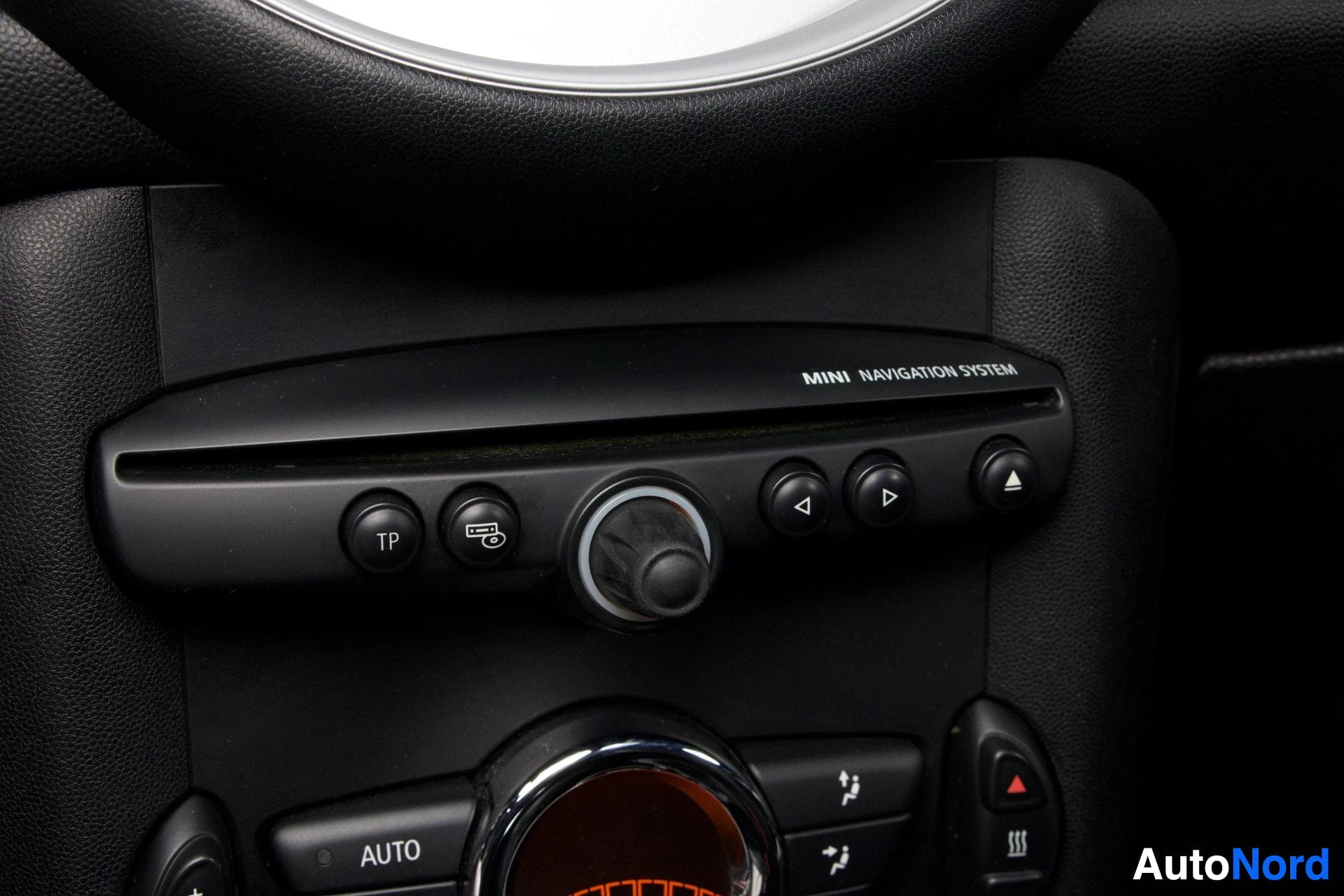 Yksityiskohta: Mini Cooperin roolinimikkeen ja navigaatiojärjestelmän ohjaintoiminnot yläpuolella näkyvät päästä; tekstillä 'MINI NAVIGATION SYSTEM' merkattu ohjauspaneeli, jo