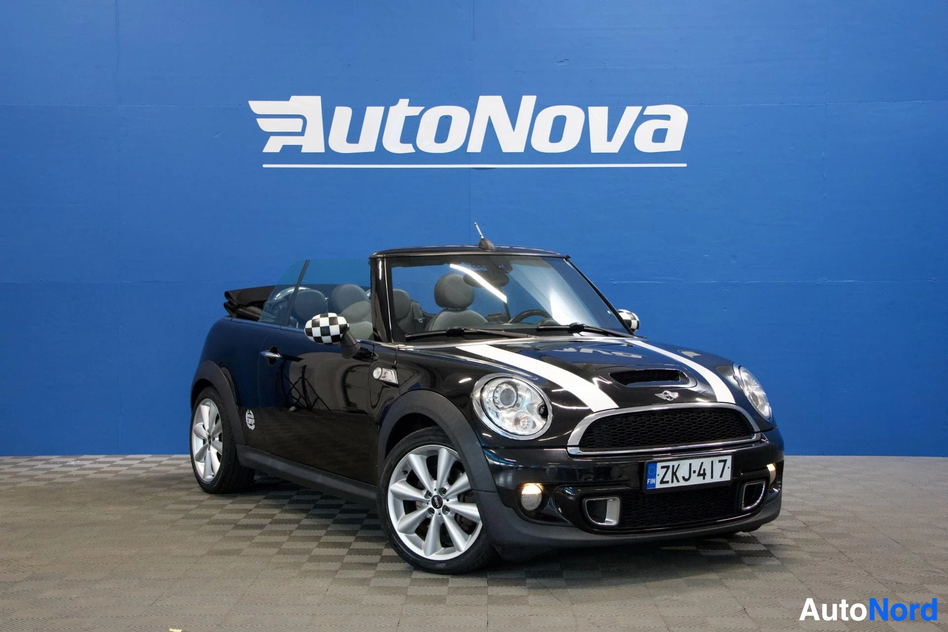 Etuviisto, nokka vasemmalle: Musta Mini Cooper avoauto edestä kolmen neljänneksen kulmasta, kone nokka vasemmalle, valkoisen ja mustien vaakakaistojen kunnailla, valkoiset alumiini