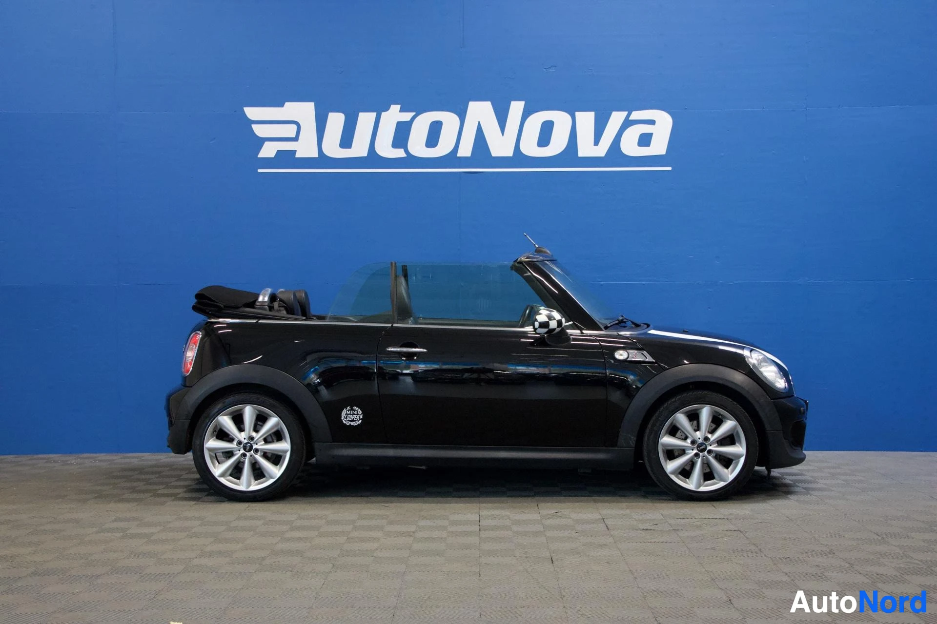 Sivulta: Musta Mini Cooper avoauto sivusta, katto avattu osittain, valkoiset alumiini-vanteet, sininen tausta