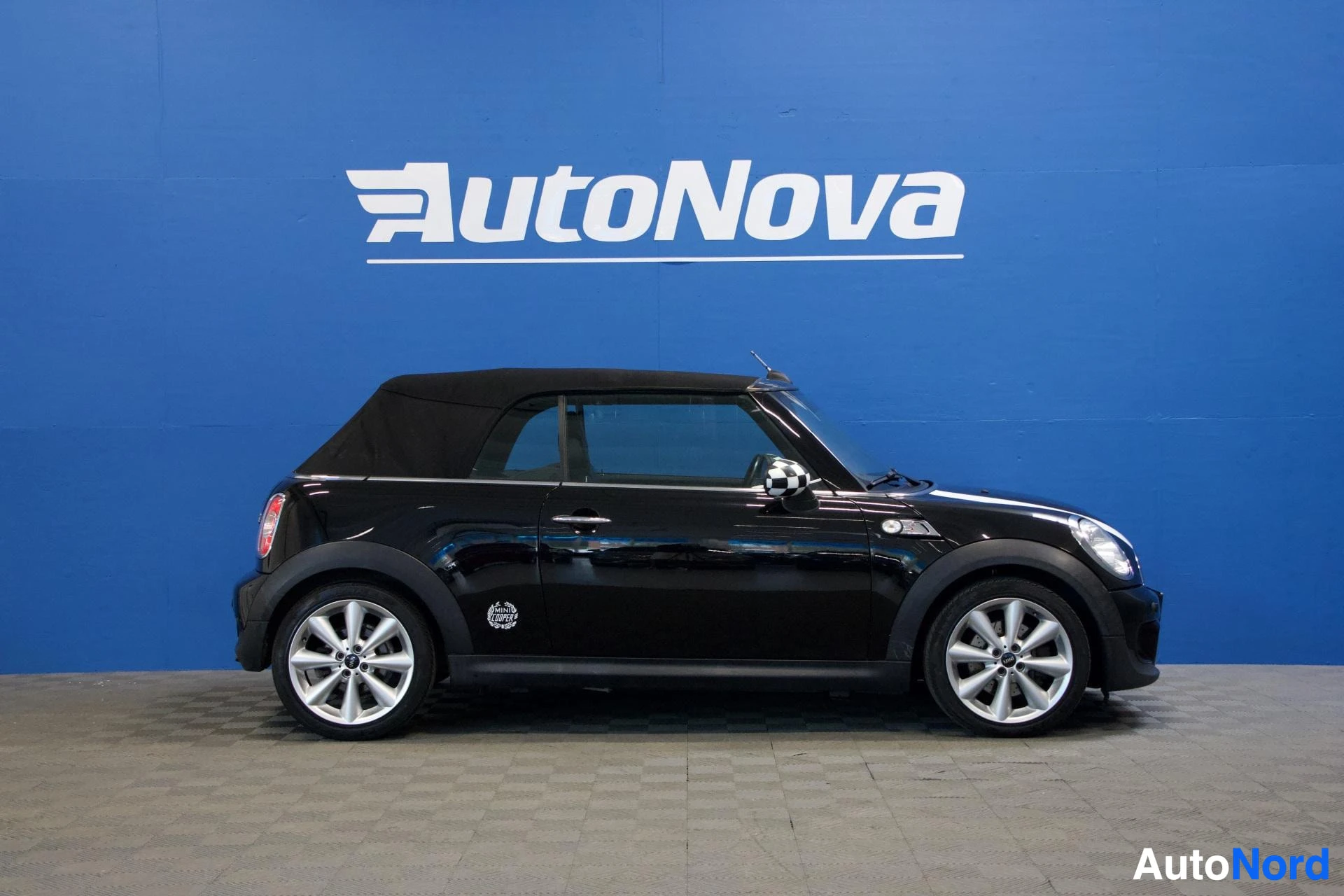 Sivulta: Musta Mini Cooper avoauto sivusta, katto suljettu, valkoiset alumiini-vanteet, sininen tausta