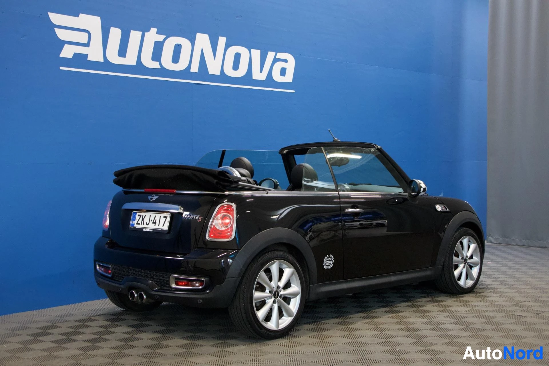 Takaviisto: Musta Mini Cooper avoauto takaa kolmen neljänneksen kulmasta, kone nokka oikealle, valkoiset alumiini-vanteet, auton katto avattu, rekisterikilpi näkyy, sininen