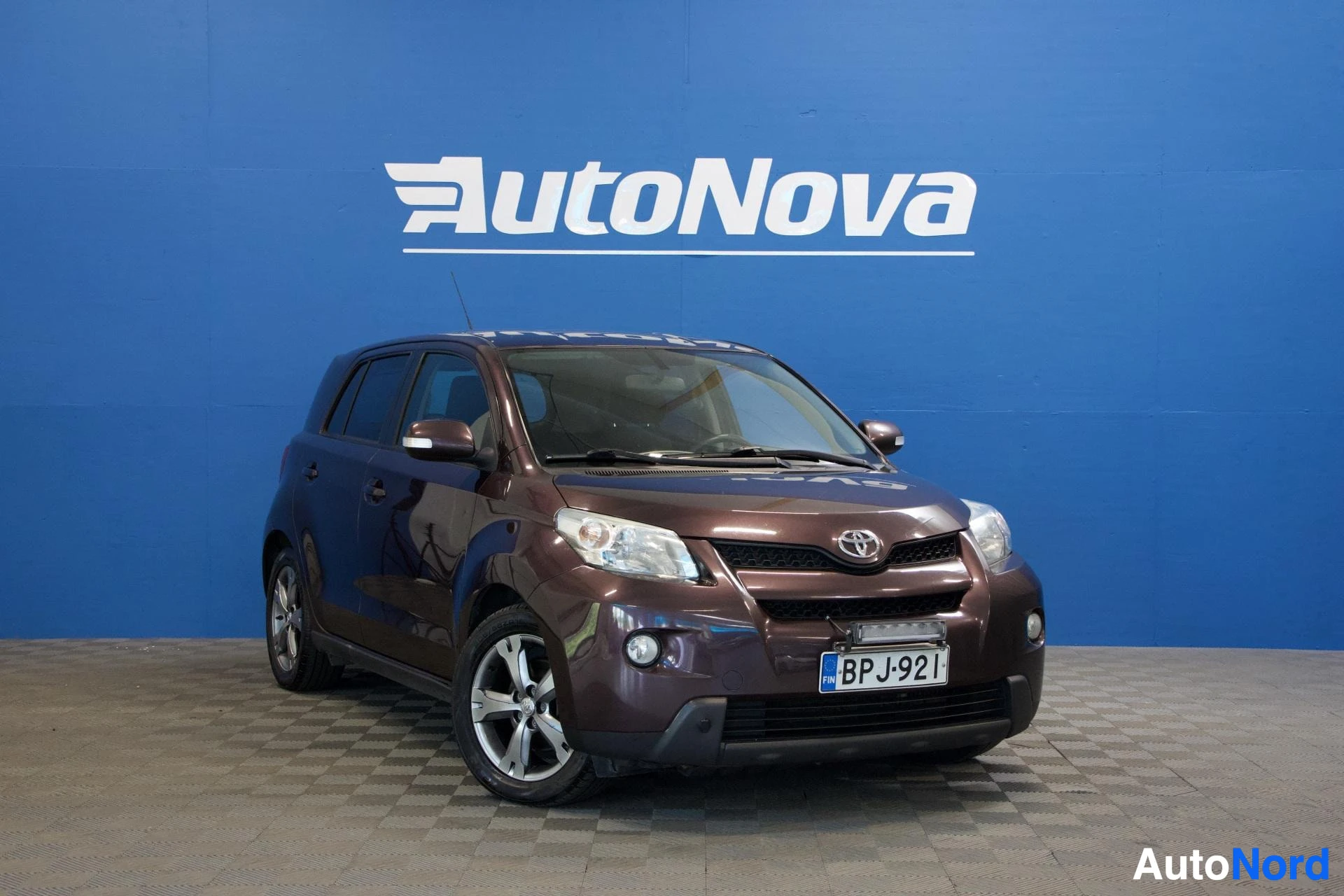 Edestä: Ruskea Toyota Urban Cruiser on pysäköity autonäyttelyn tiloihin AutoNova-logolla varustetun sinisen seinän eteen. Ajoneuvon etuosa on näkyvissä, ja rekisterikil