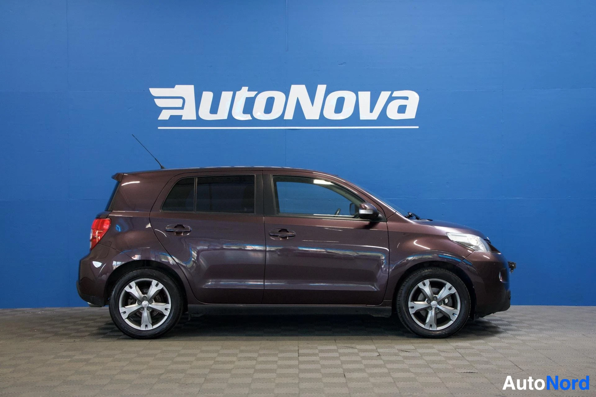 Sivulta: Ruskea Toyota Urban Cruiser on pysäköity autonäyttelyn tiloihin AutoNova-logolla varustetun sinisen seinän eteen. Ajoneuvon vasen kylki on näkyvissä. Autossa on