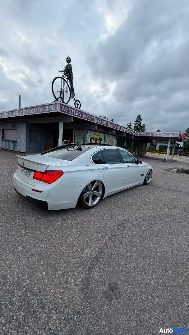 Takaviisto: Valkoinen BMW-sedan oikealta sivulta taaksepäin, pysäköitynä asfalttialueelle. Oikea sivu ja takaosa näkyvät. Rekisterikilpi näkyy. Taustalla rakennuksella polk