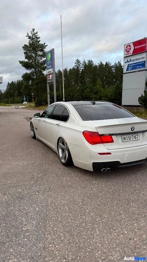Takaviisto: Valkoinen BMW-luksussarja-sedan pysäköitynä asvalttialueelle, sivunäkymä oikealta takaa, rekisterikilvessä NTU751, taustalla myymälöitä ja metsää.
