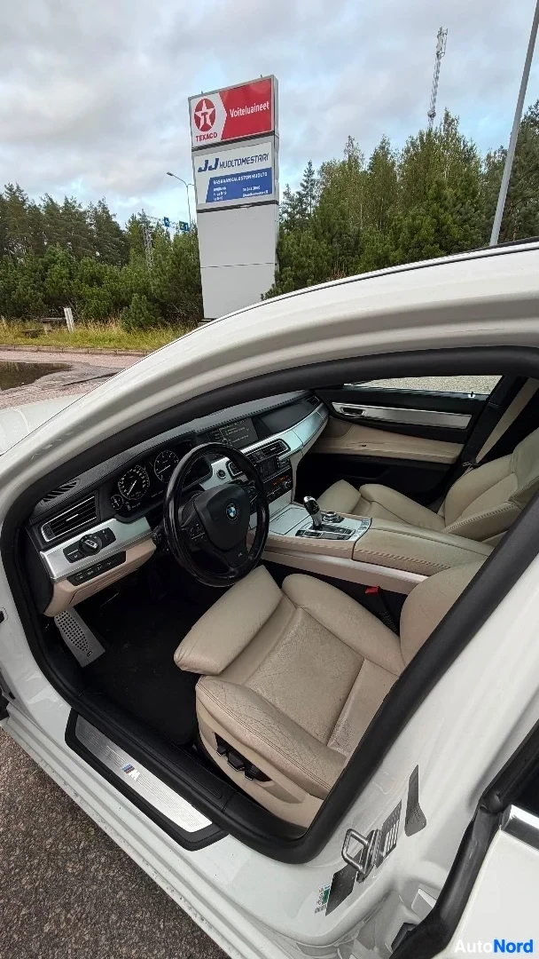 Etuistuimet: Auton sisänäkymä avoimesta kuljettajan ovesta, näkyy vaalean väriset nahkaistuin, mustatekstuuroitu kojelauta BMW-logolla ja ohjausrata, taustalla huoltoaseman