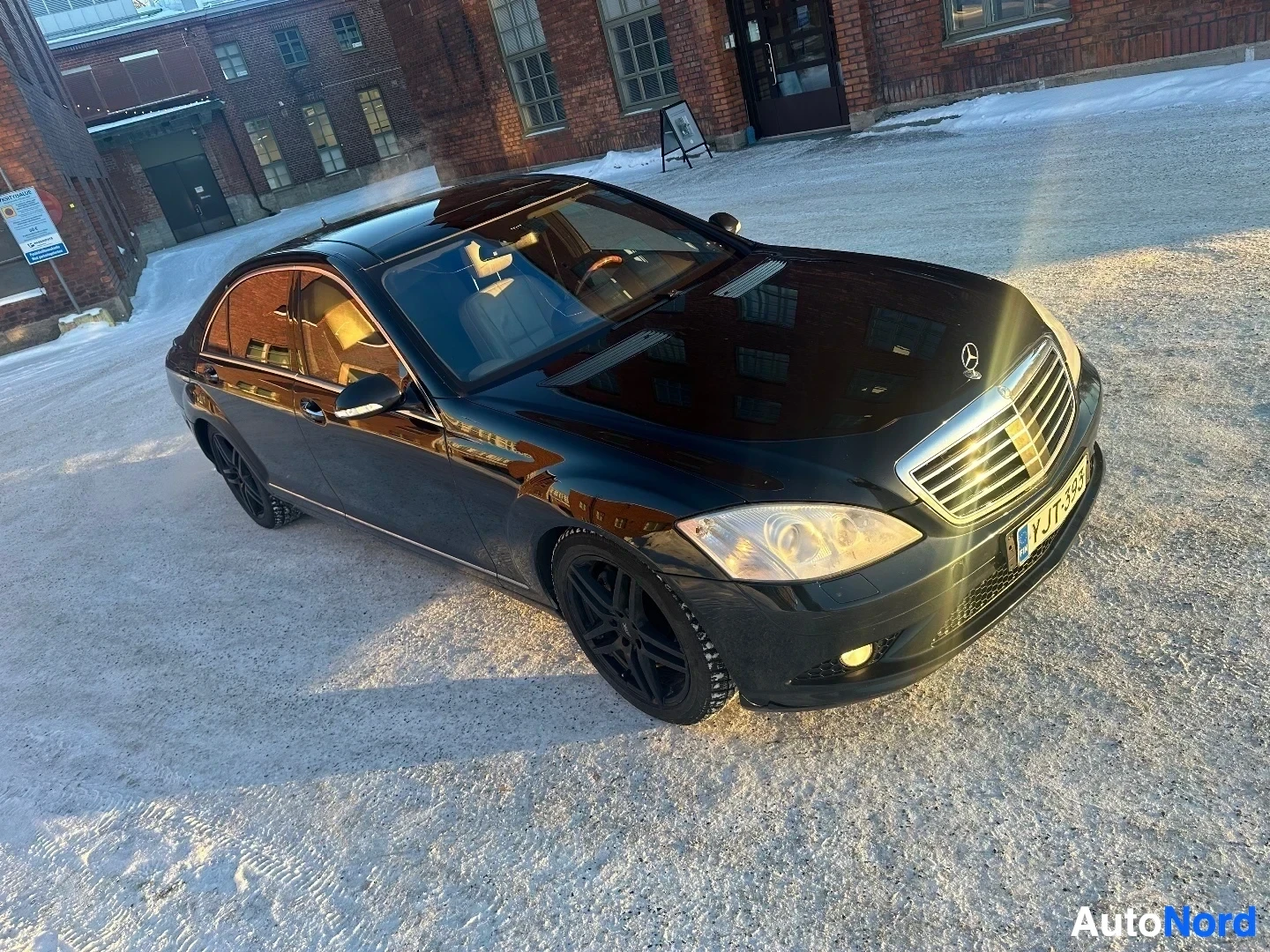 Etuviisto, nokka vasemmalle: Musta Mercedes-Benz sedan, näkymä etuviistosta vasemmalta, pysäköity tiilirakennus‍taustalla olevalle alueelle