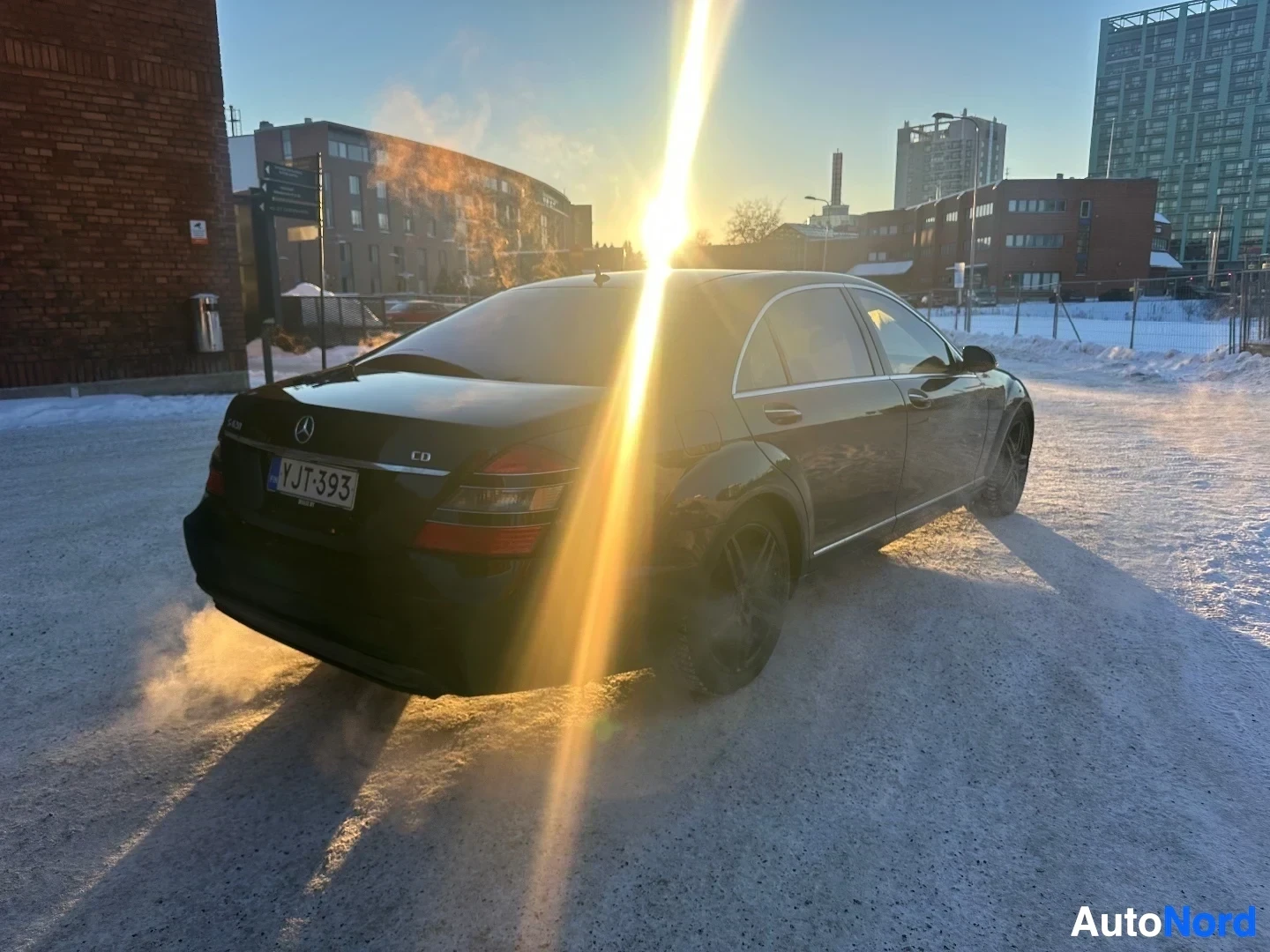 Takaviisto: Musta Mercedes-Benz sedan takaviistosta, pysäköity lumiselle alueelle, aurinko takana aiheuttaa hehkua