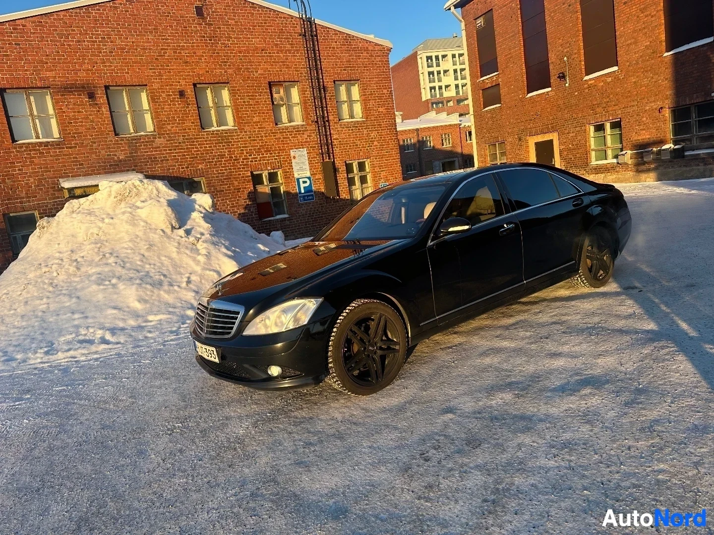 Sivulta: Musta Mercedes-Benz sedan sivusta oikealle, pysäköity tiilirakennus‍taustalla olevalla alueella, aurinko vasemmalta