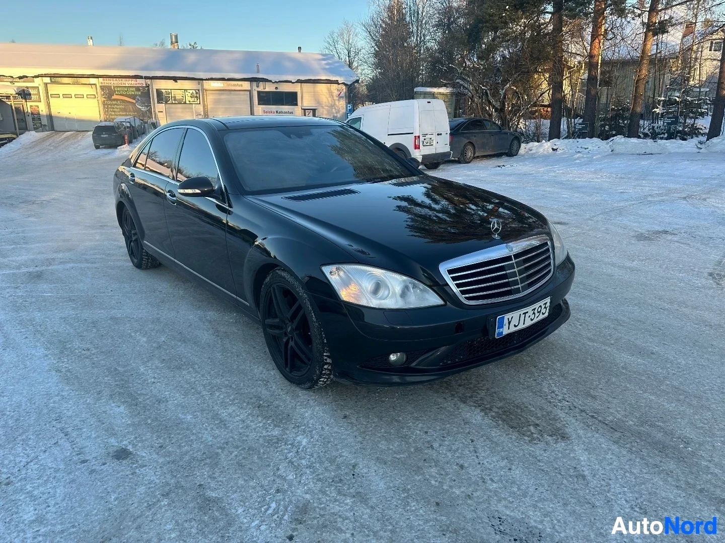 Etuviisto, nokka oikealle: Musta Mercedes-Benz S-luokan sedan edestä kolmelta neljäsosalta katsottuna, aurinko nouseva taustalla
