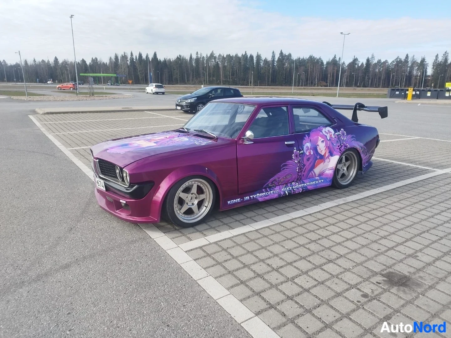 Sivulta: Violetti auto parkkipaikalla sivusta katsottuna; näkyvät kolme valaisinta etuosassa ja mustavalkoinen rekisterkilpi