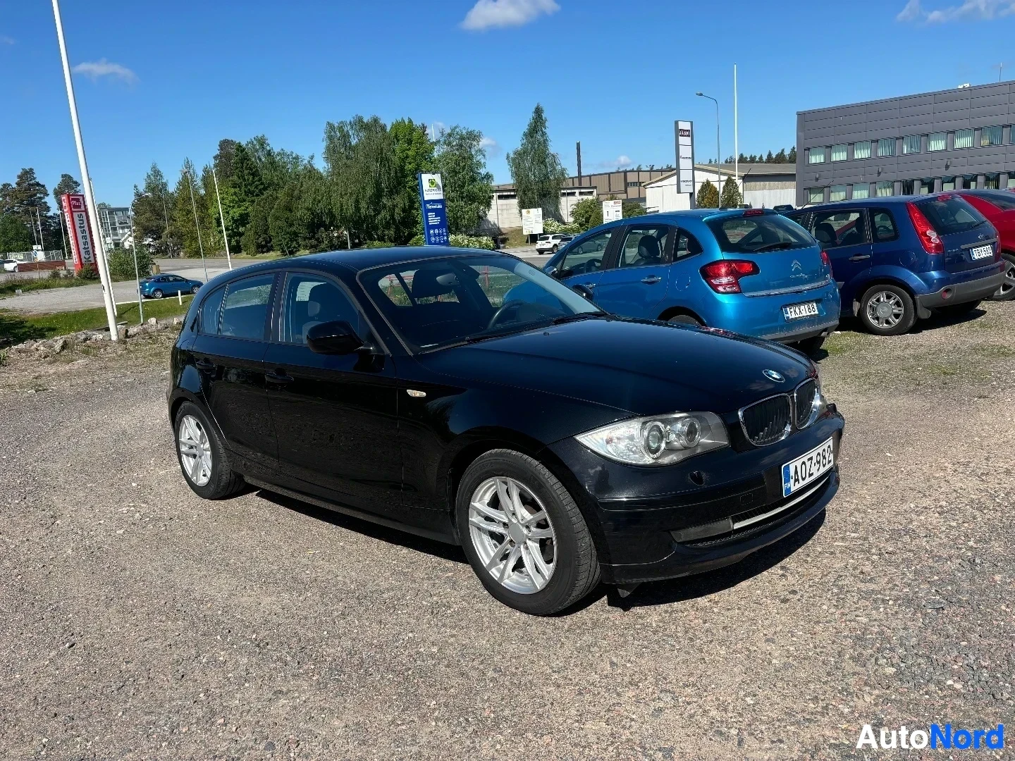 Etuviisto, nokka vasemmalle: Musta BMW 116i kolmiovi pysäköitynä autojen jälleenmyyjän pihalla, koko ajoneuvo näkyvissä, kuvattu vasemmalta etupuolelta.