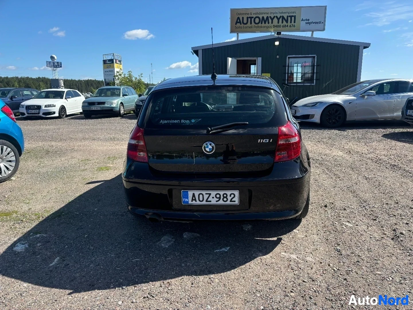Takaa: Musta BMW 116i takaa kuvattuna, näkyvissä takaosassa teksti '116i' ja rekisterikyltti, tausta autokauppias.