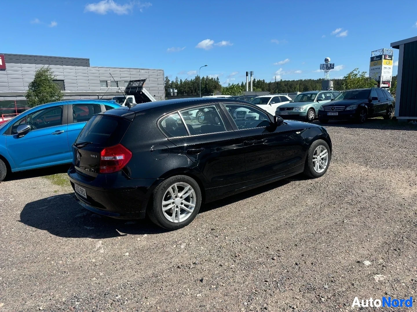 Takaviisto: Musta BMW 116i Business -malli pysäköitynä autoliikeellä, kuvattu sivulta taakse päin. Kuulta näkyy avoin konepelti ja valkoisen väriset vanteet. Taustalla muit