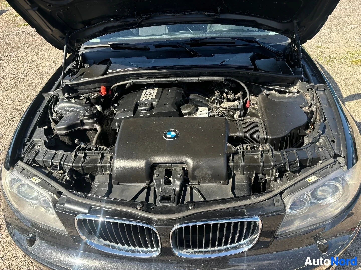 Moottoritila: BMW:n konepelti on täysin auki. Näkyy musta moottori BMW-logolla, ilmansuodatinlaatikko, ja erilaisia verkkomaisia moottoriin liittyviä osia. Pääkoneen perälle
