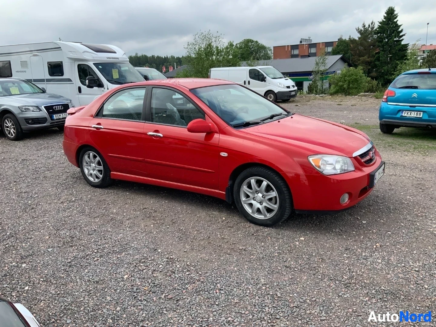 Sivulta: Punainen Kia Cerato nelioven sedan sivulla seisten, oikea sivu näkyvissä, auton nosto edessä ja vasemmalla.