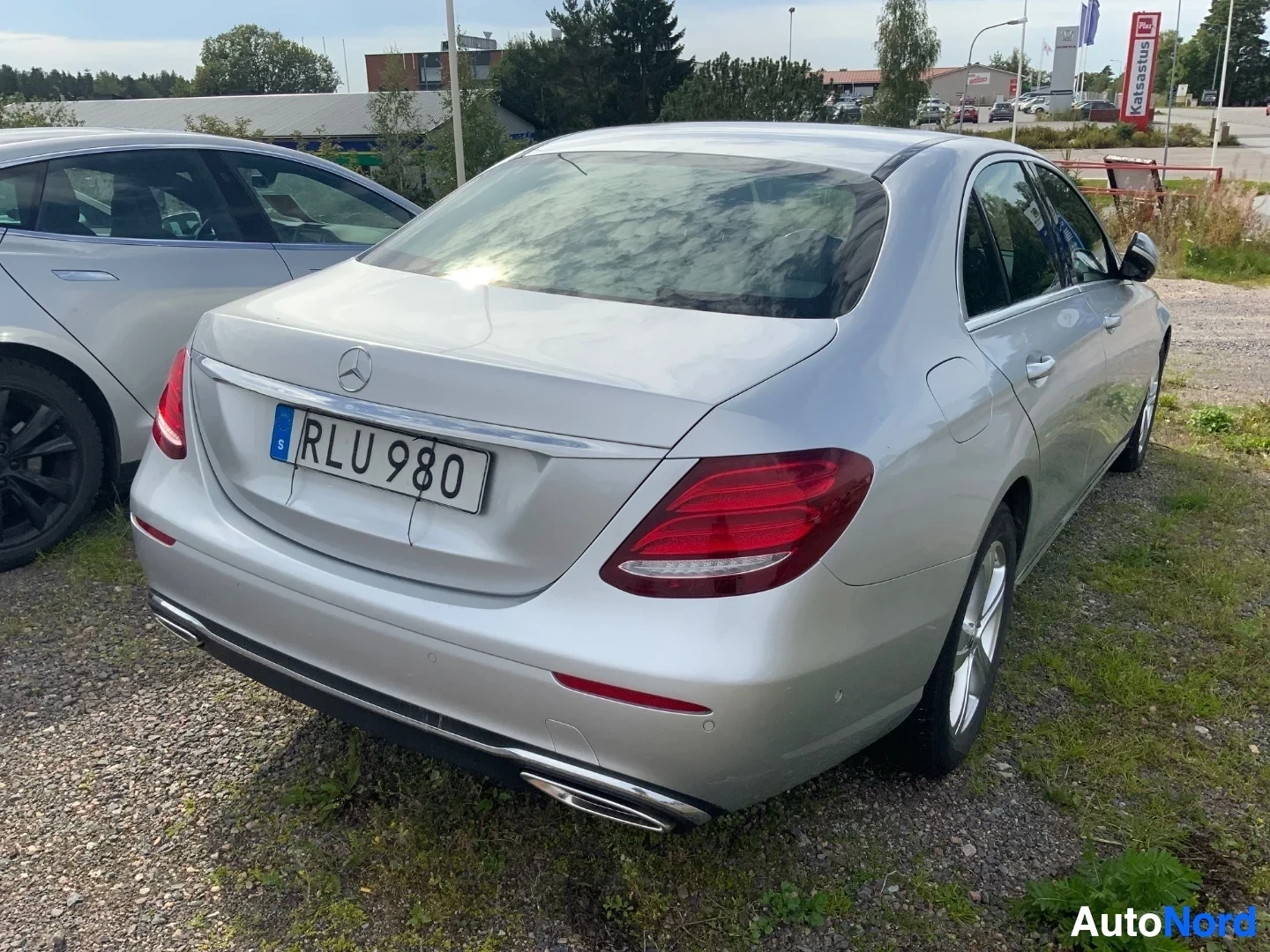 Takaa: Hopeanvärinen Mercedes-Benz sedan, takaa kuvattuna. Rekisterikilvessä RLU 980. Auto seisoo sorareunuksella, taustalla kauppakeskusympäristö.