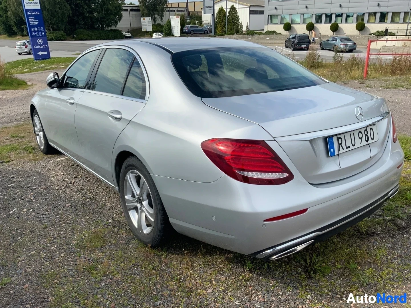 Takaviisto: Hopeanvärinen Mercedes-Benz sedan, oikealta sivulta ja takaosasta kuvattuna. Rekisterikilvessä RLU 980. Auto seisoo sorareunuksella, taustalla kauppakeskusympär