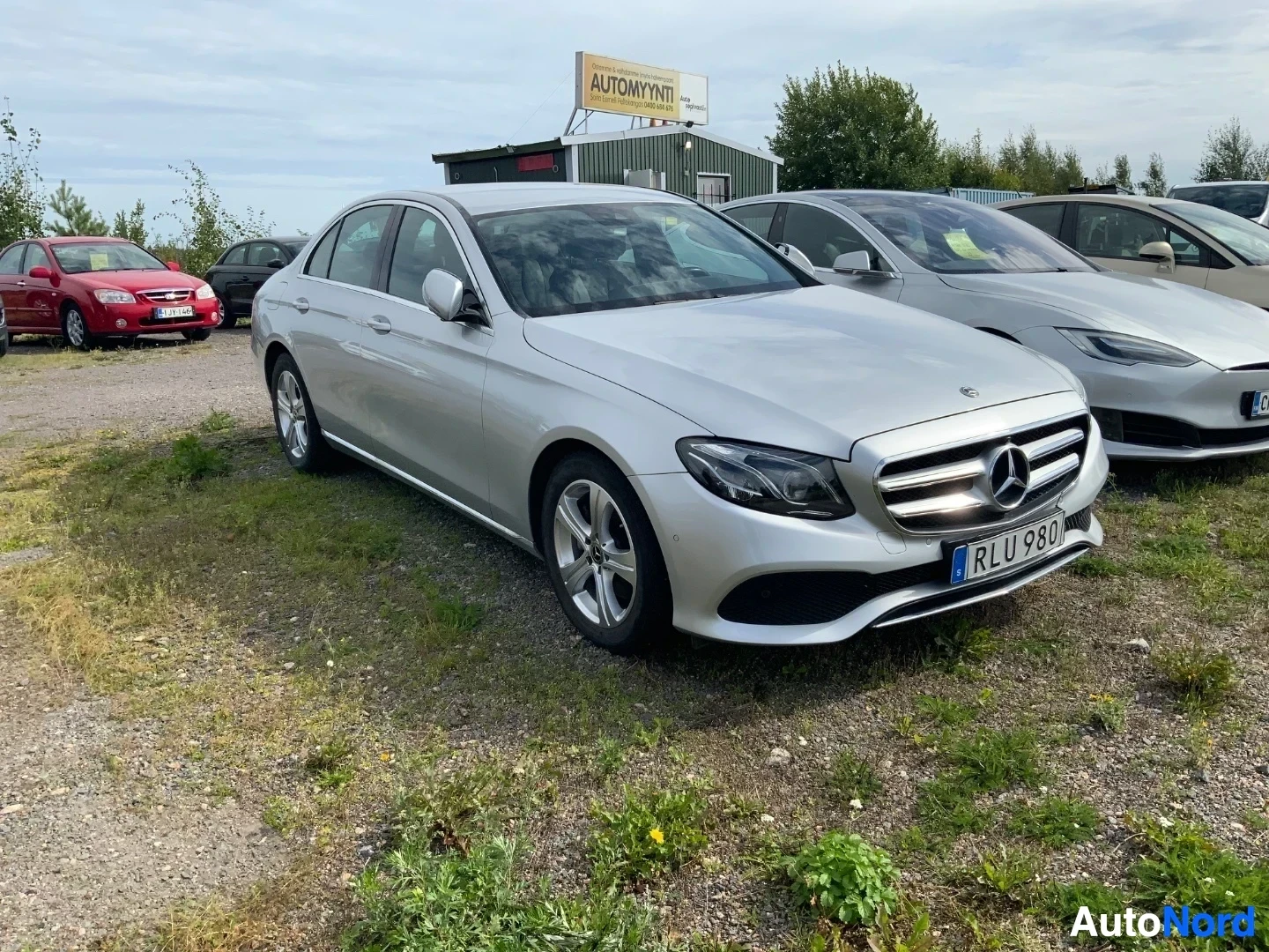 Etuviisto, nokka vasemmalle: Valkoinen Mercedes-Benz E-sarja sedan, kuvattu kolmesta neljäsosasta etupuolelta vasemmalta, kaupallialla autojen myyntialueella.