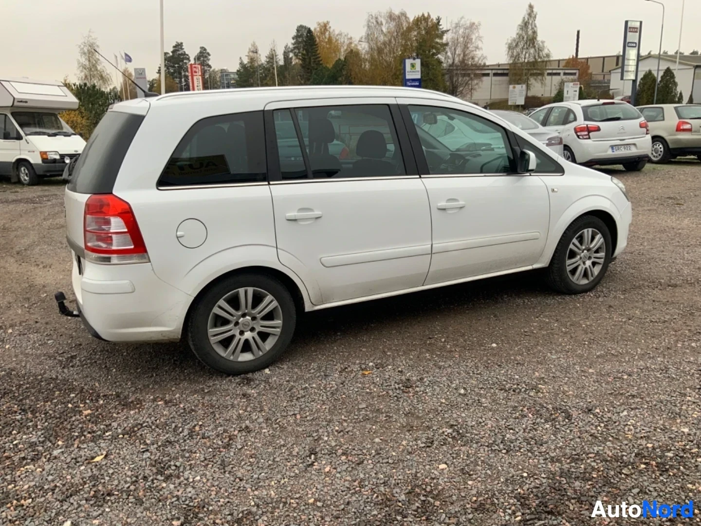 Sivulta: Valkoinen Opel Zafira -tila-auto sivukuvassa oikealta. Näkyvissä sivuprofiili, ovet ja punaiset takavalot. Rekisterilevy näkyvissä