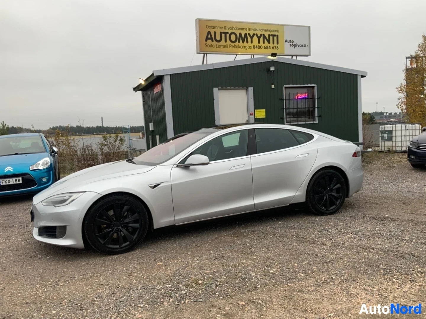 Sivulta: Valkoinen Tesla-sedan kuvattuna sivusta autojen myyntiliikkeen pihalla. Auton profiilikuvassa näkyvät mustat aluevanteet, tummennuslasi ja kokonaismuoto.