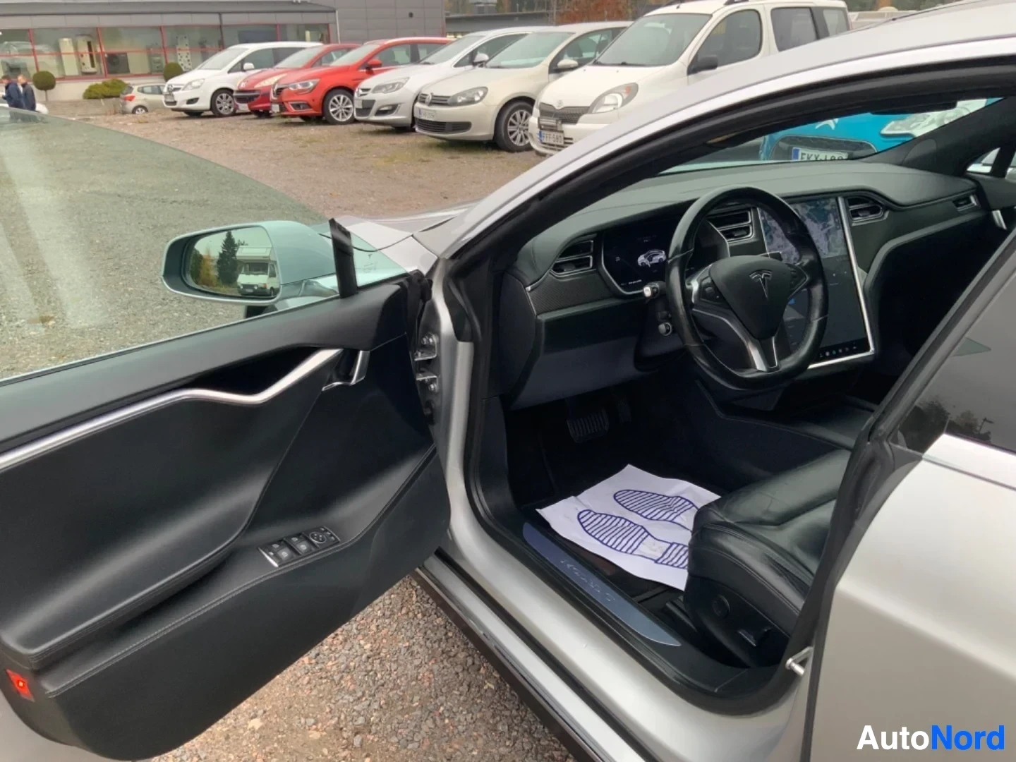 Ohjaamo: Ajoneuvon sisustö sivusta katsottuna. Mustat urheilu-istuimet, Tesla-ohjauspyörä, kosketusnäyttö näkyvät. Ovet auki, paperit näkyvät istuimella.