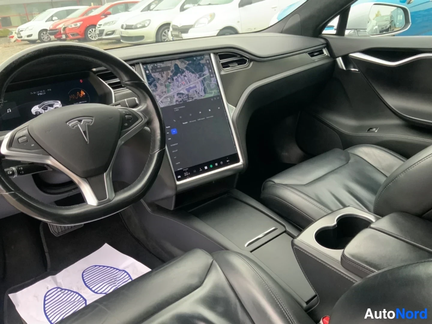 Ohjaamo: Auton sisäkuva etulta: mustavalkoinen ohjaamo Tesla-logolla varustetulla ohjauspyörällä, suurella kosketusnäytöllä navigointikartalla, nahkaistuimilla ja monios