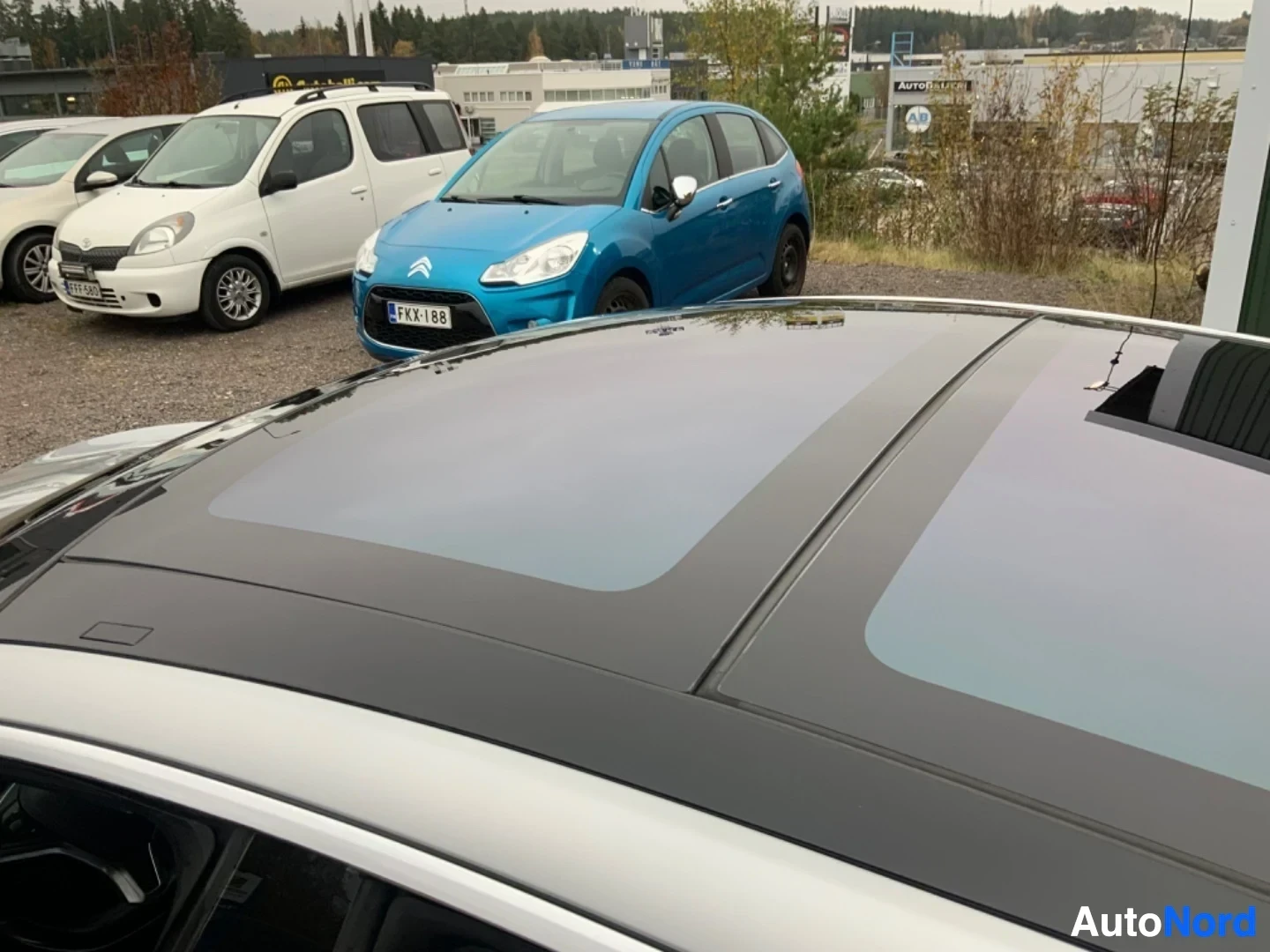 Muu: Valkoisen sedan-auton katto ylhäältä kuvattuna, näkyy panoraamisen auringonkattoaukko ja etäisyydessä pysäköityjen autojen kattoja.