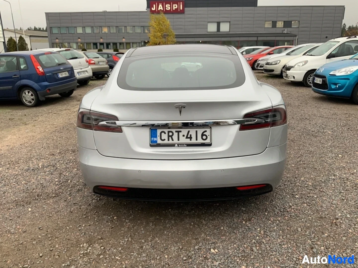 Takaa: Hopeinen Tesla-sedan näkyy takaapäin autokauppiaiden pihalla. Rekisterilukuna