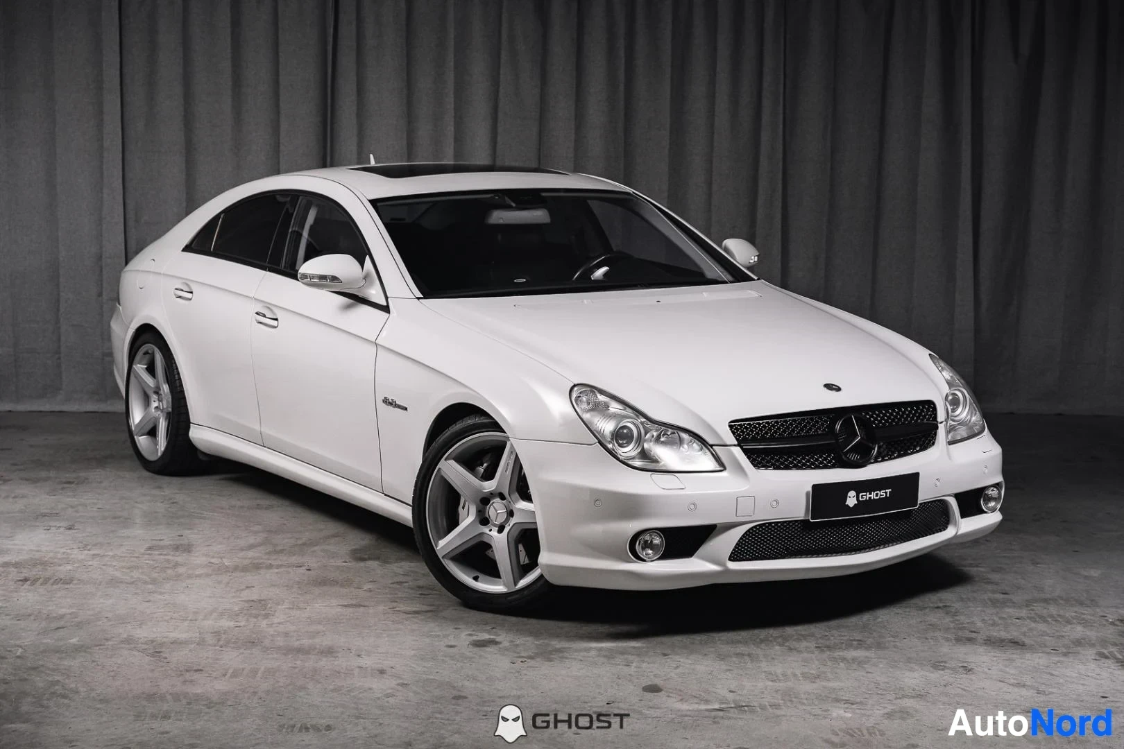 Etuviisto, nokka oikealle: Valkoinen Mercedes-Benz CLS 63 AMG, koko ajoneuvo näkyvissä, hopeanväriset AMG-vanteet, studiotausta