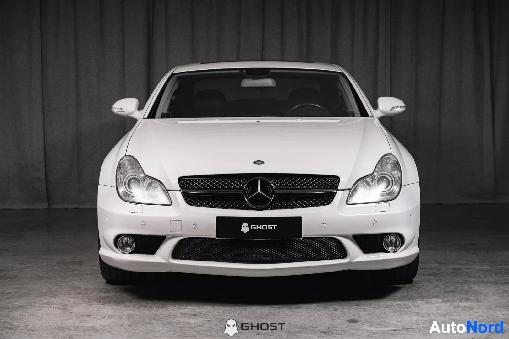 Edestä: Valkoinen Mercedes-Benz CLS 63 AMG, koko ajoneuvo näkyvissä, ajovalot päällä, studiotausta