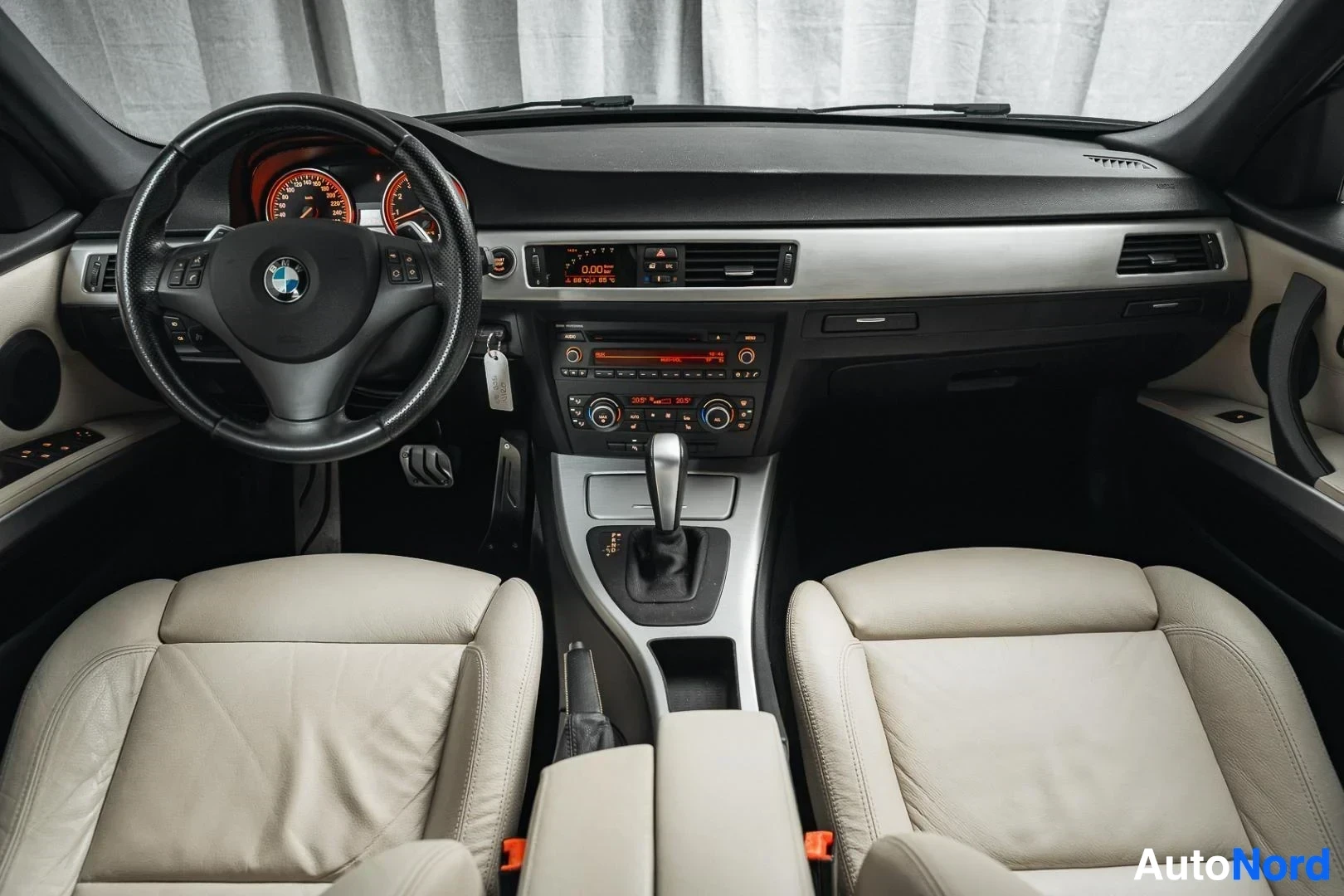 Ohjaamo: Musta BMW 335i xDrive E91 Touring, ohjauspyörä, mittaristo ja keskikonsoli takaa kuvattuna, beige nahkaverhoilu