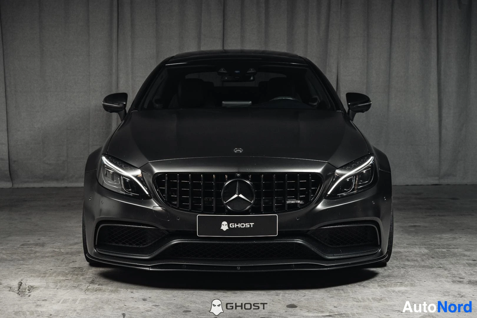 Edestä: Mattamusta Mercedes-Benz C 63 AMG coupé, koko ajoneuvo näkyvissä, ajovalot päällä, studiotausta