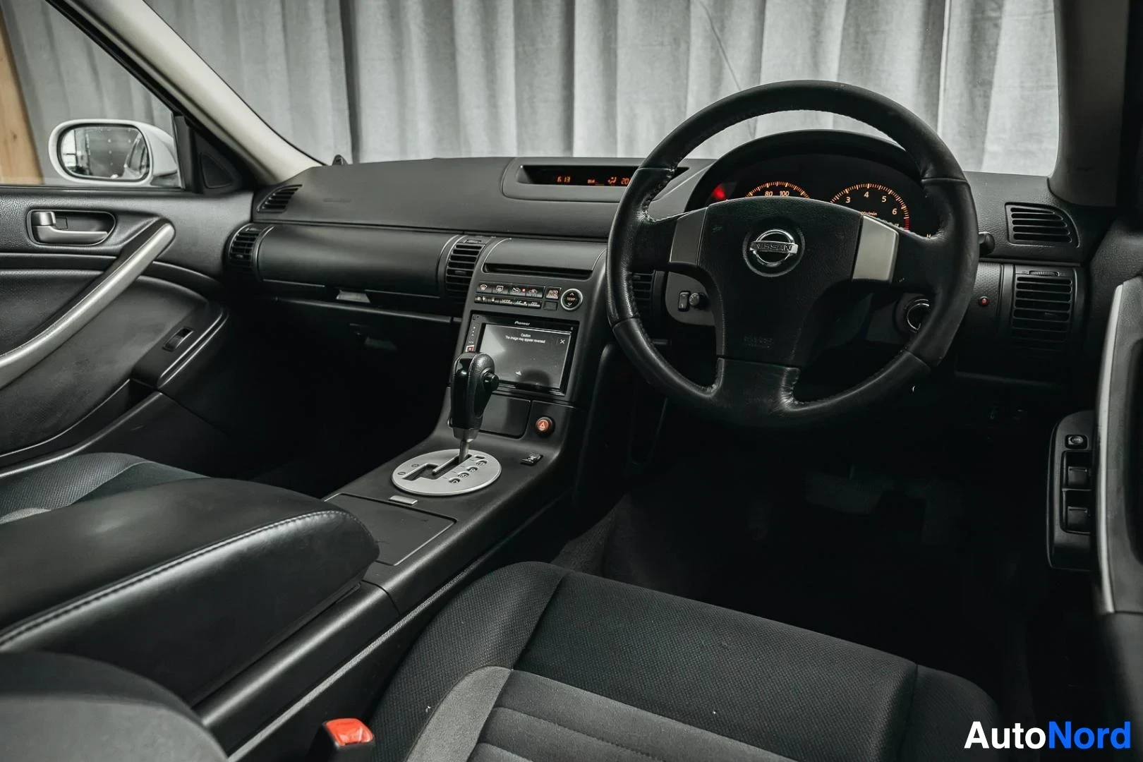 Ohjaamo: Valkoinen Nissan Stagea, kojelauta, ohjauspyörä ja keskikonsoli matkustajan puolelta