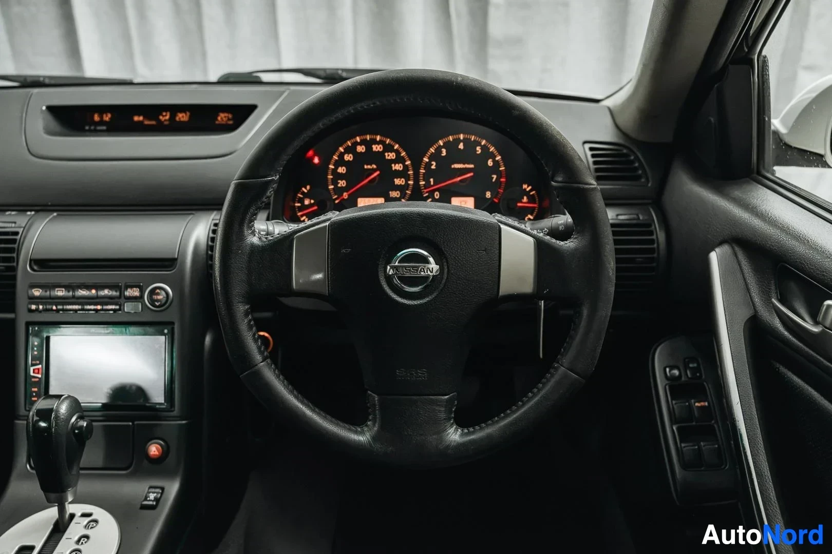 Ohjaamo: Valkoinen Nissan Stagea, ohjauspyörä, mittaristo ja keskinäyttö kuljettajan puolelta