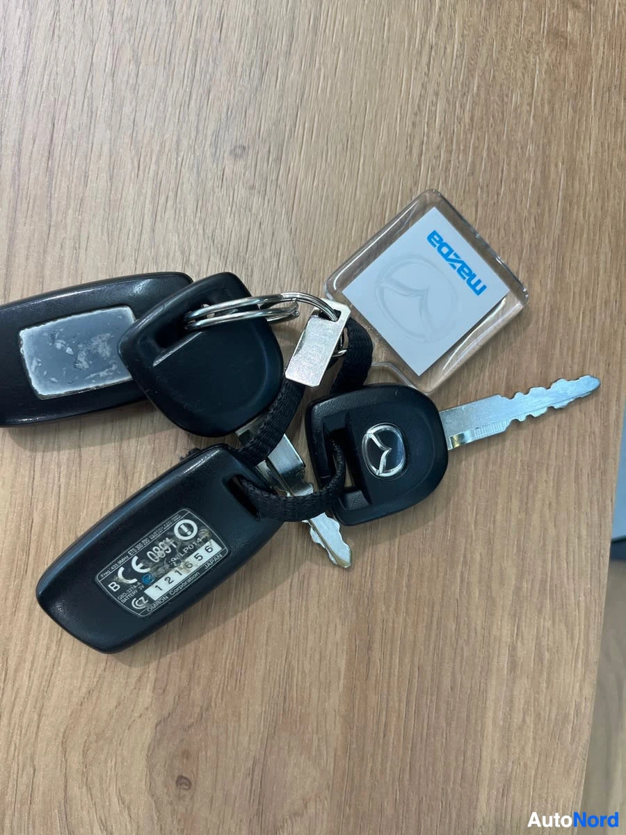 Yksityiskohta: Kaksi Mazda-avaimenperää, joissa on avaimet, puupöydällä. Toinen avaimenperä on läpinäkyvä ja siinä on Mazda-logo, toinen on musta kaukosäädinavain, jossa on te