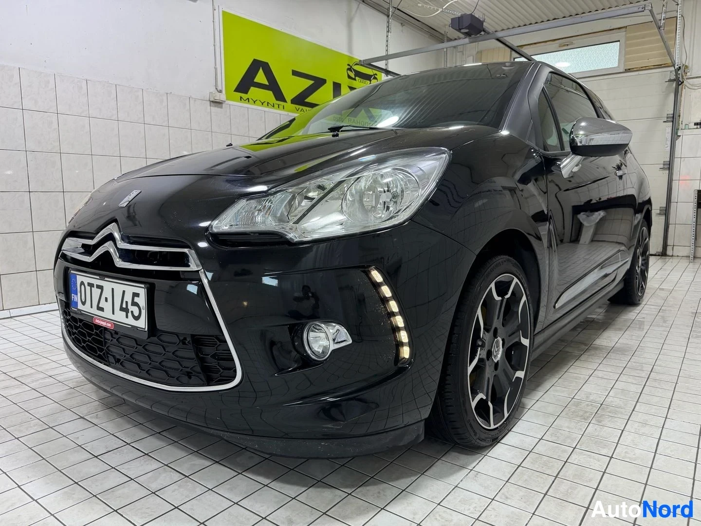 Edestä: Musta Citroën DS3 pysäköitynä halliin, etunäkymä. Rekisterikilpi näkyy. Autolla on LED-ajovalot ja valkoiset vanteet. Taustalla keltainen AZIZ-kyltti