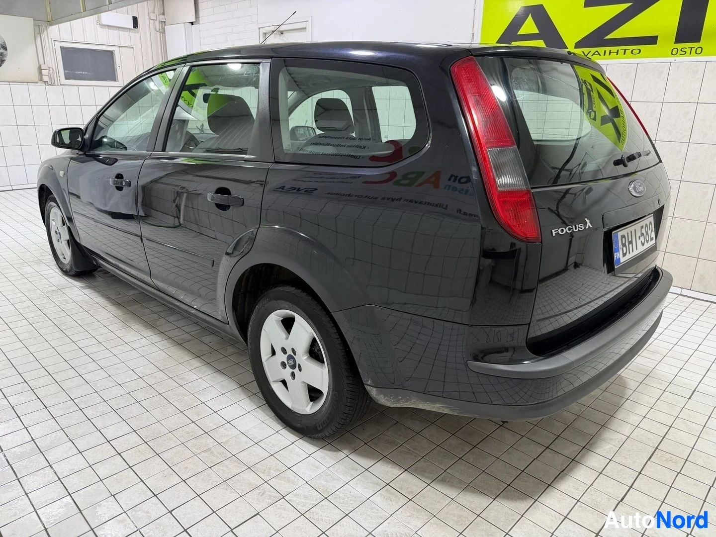 Takaviisto: Musta perhonauto näkymä takaa-sivulta oikealta, oven ollessa auki, sisätiloissa keltaisen Azix-kyltin edessä