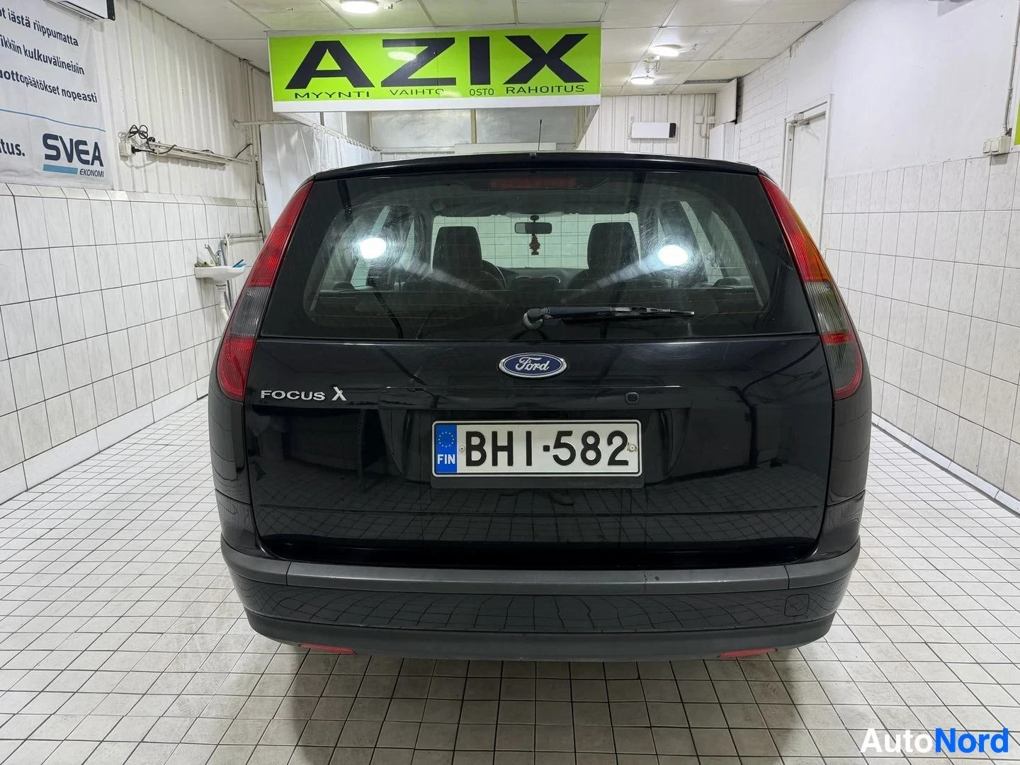 Takaa: Musta auto takaapäin, takalasi näkyvissä, Ford Focus X -merkintä ja sininen rekisterikylpi takaosassa, punaiset jarrovalot